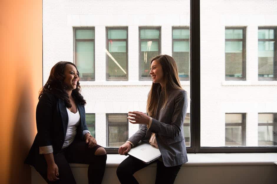 Two women sitting on a window sill talking.