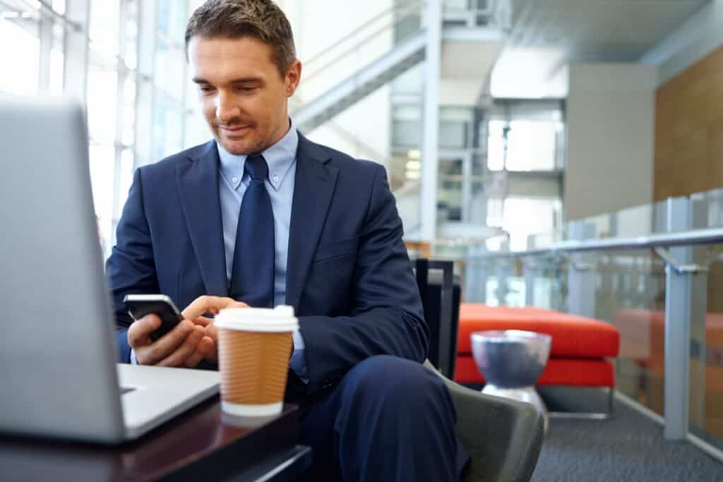 A man in a suit sitting at a desk with a cup of coffee and a cell phone.